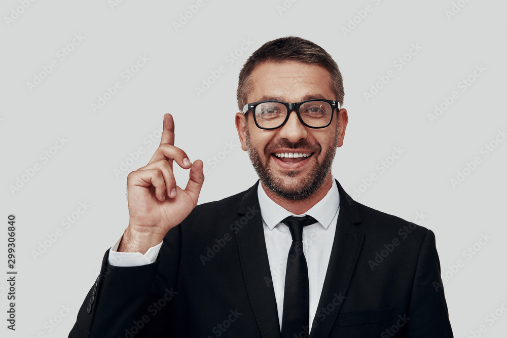 Charming young man in full suit smiling and pointing copy space while standing against grey background Charming young man in full suit smiling and pointing copy space while standing against grey background