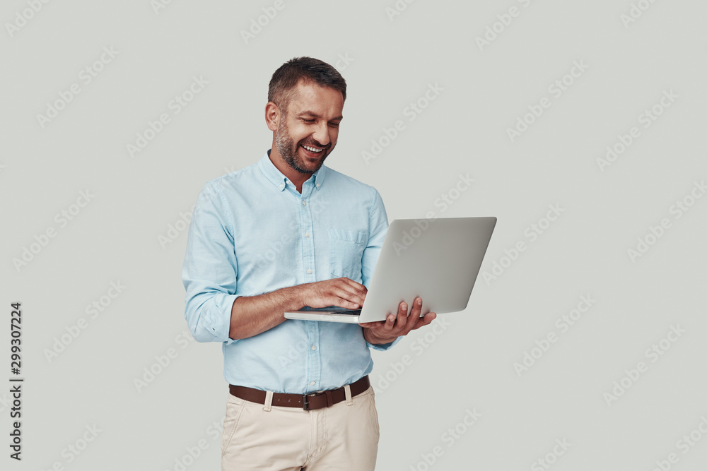 Handsome young man using laptop and smiling while standing against grey background Handsome young man using laptop and smiling while standing against grey background