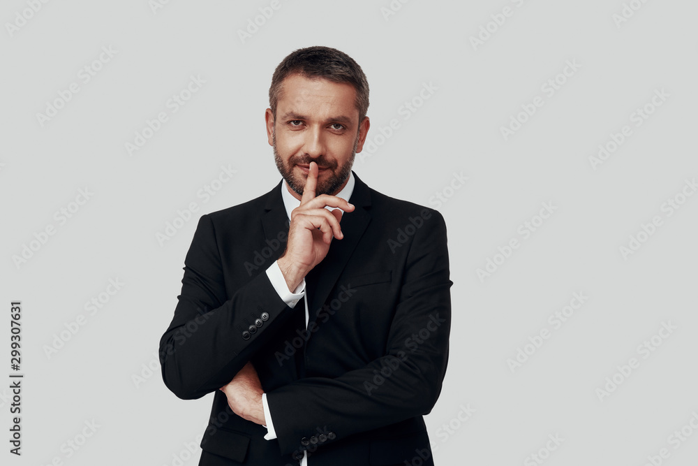 Charming young man in full suit keeping finger on lips while standing against grey background Charming young man in full suit keeping finger on lips while standing against grey background