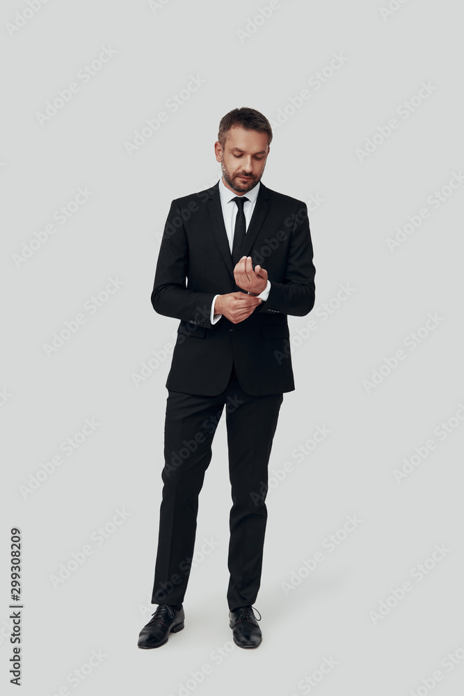 Full length of charming young man in full suit adjusting sleeve while standing against grey background Full length of charming young man in full suit adjusting sleeve while standing against grey background