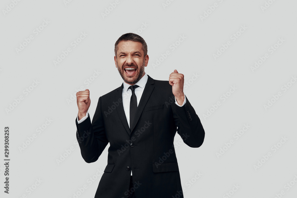 Charming young man in full suit cheering while standing against grey background Charming young man in full suit cheering while standing against grey background
