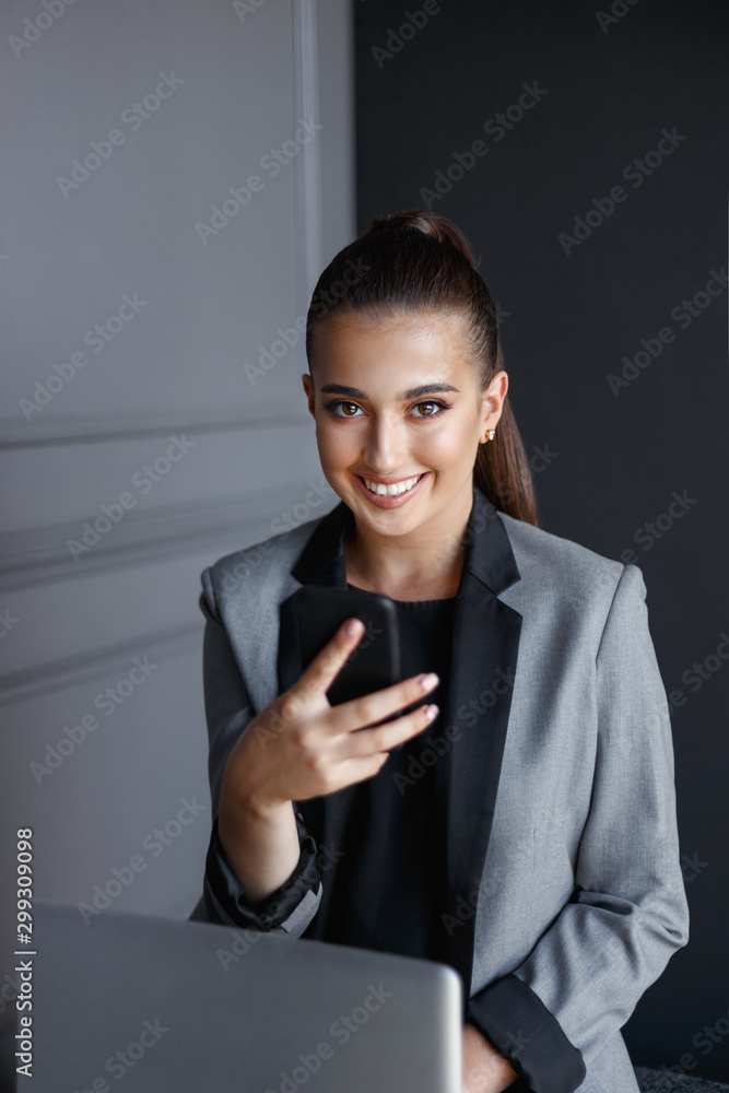 Cheerful girl with adorable smiling, in grey and black suit, working on laptop, using smartphone over grey background. Cheerful girl with adorable smiling, in grey and black suit, working on laptop, using smartphone over grey background.
