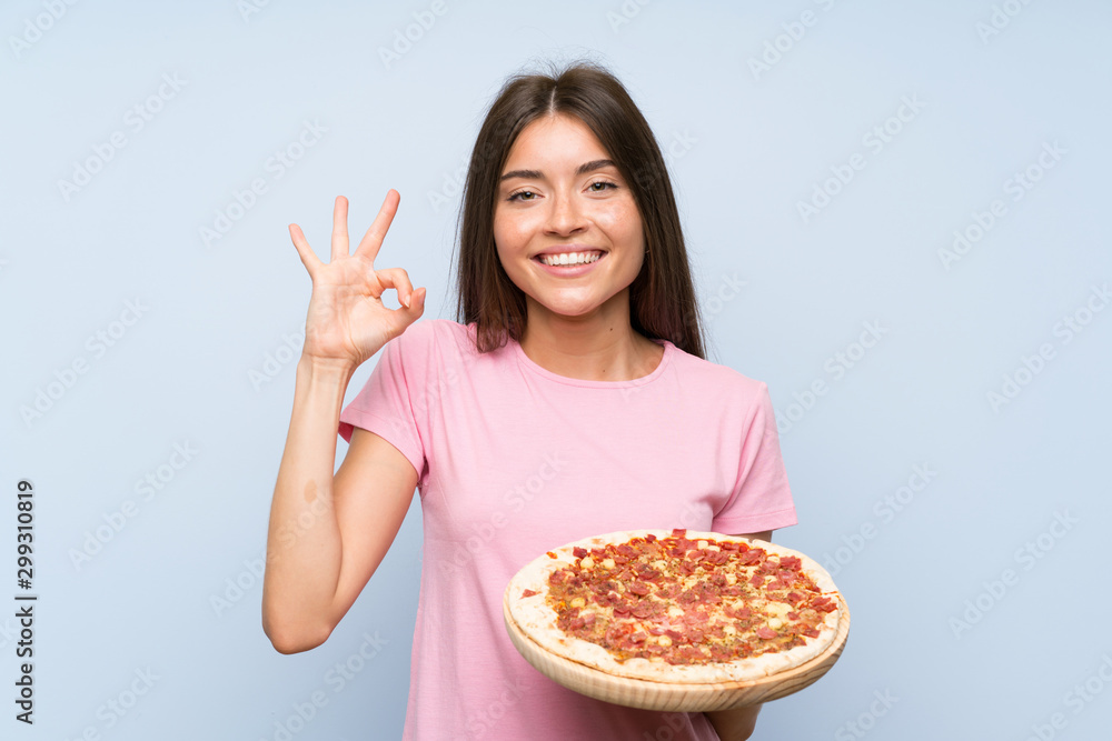 Pretty young girl holding a pizza over isolated blue wall showing ok sign with fingers