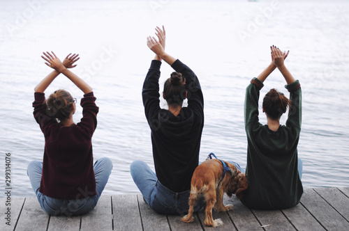  Three young women doing yoga at nature. Fitness, sport, yoga and healthy lifestyle concept - group of people making yoga pose on lake pier at sunset