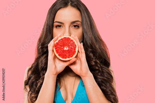 young woman in swimsuit covering face with half of grapefruit isolated on pink