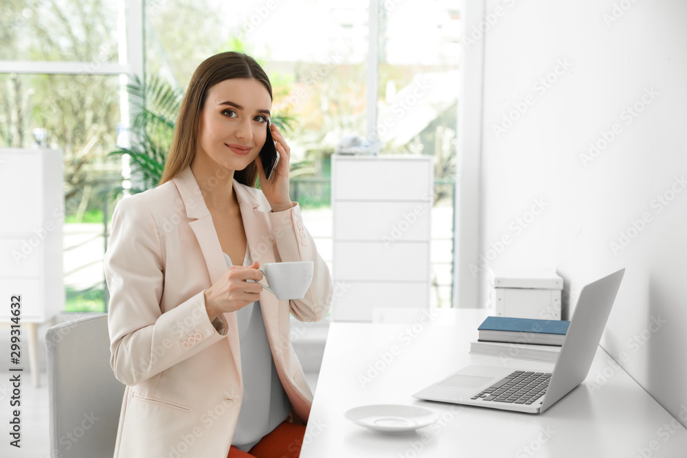 Young businesswoman talking on phone while using laptop at table in office Young businesswoman talking on phone while using laptop at table in office