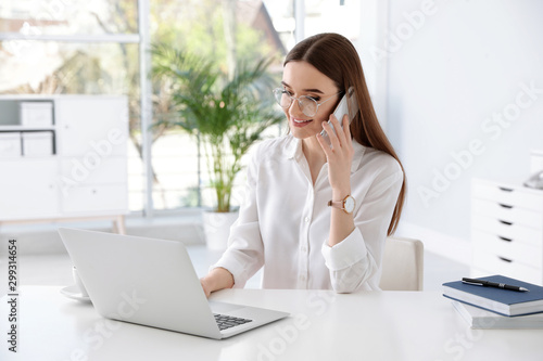 Young businesswoman talking on phone while using laptop at table in office photo