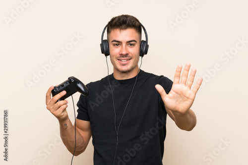 Young handsome man playing with a video game controller over isolated background saluting with hand with happy expression