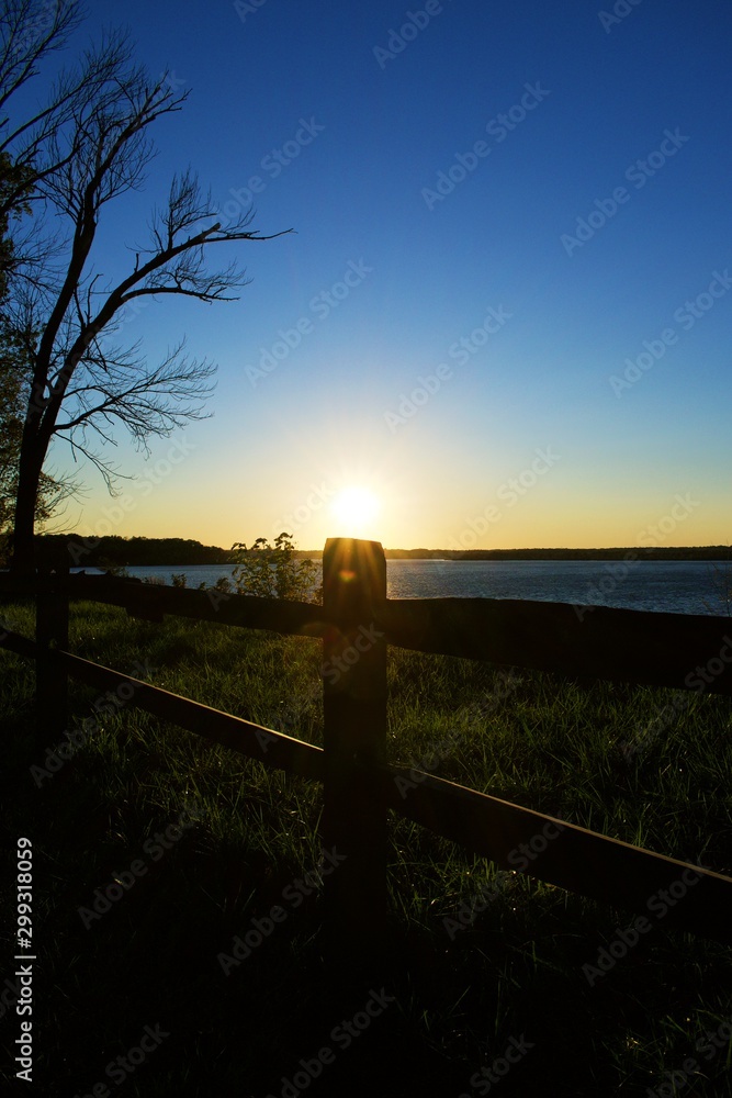 Sunset over the water with Sun resting on wood fence post.