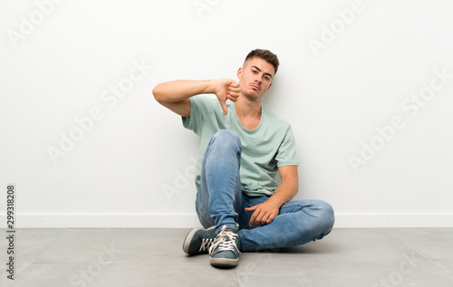 Young handsome man sitting on the floor showing thumb down sign