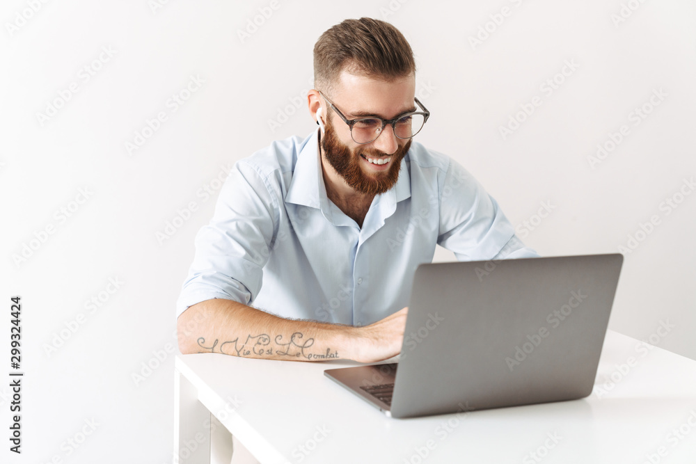 Image of man wearing eyeglasses and earbuds working on laptop in office Image of man wearing eyeglasses and earbuds working on laptop in office