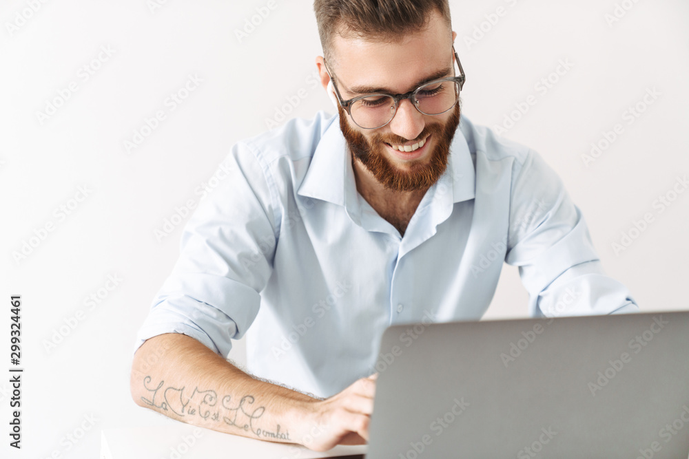 Image of man wearing eyeglasses and earbuds working on laptop in office Image of man wearing eyeglasses and earbuds working on laptop in office