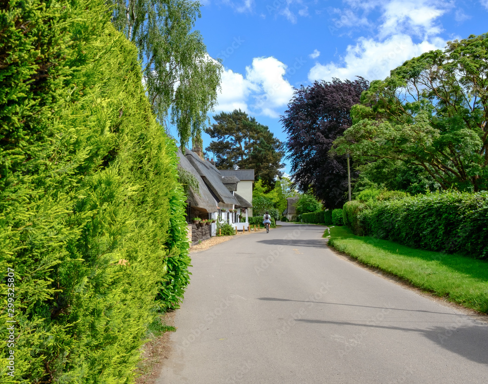 Quintessential view of a typical English village showing an empty narrow road with cottages together with a distant cyclist on a summers day.