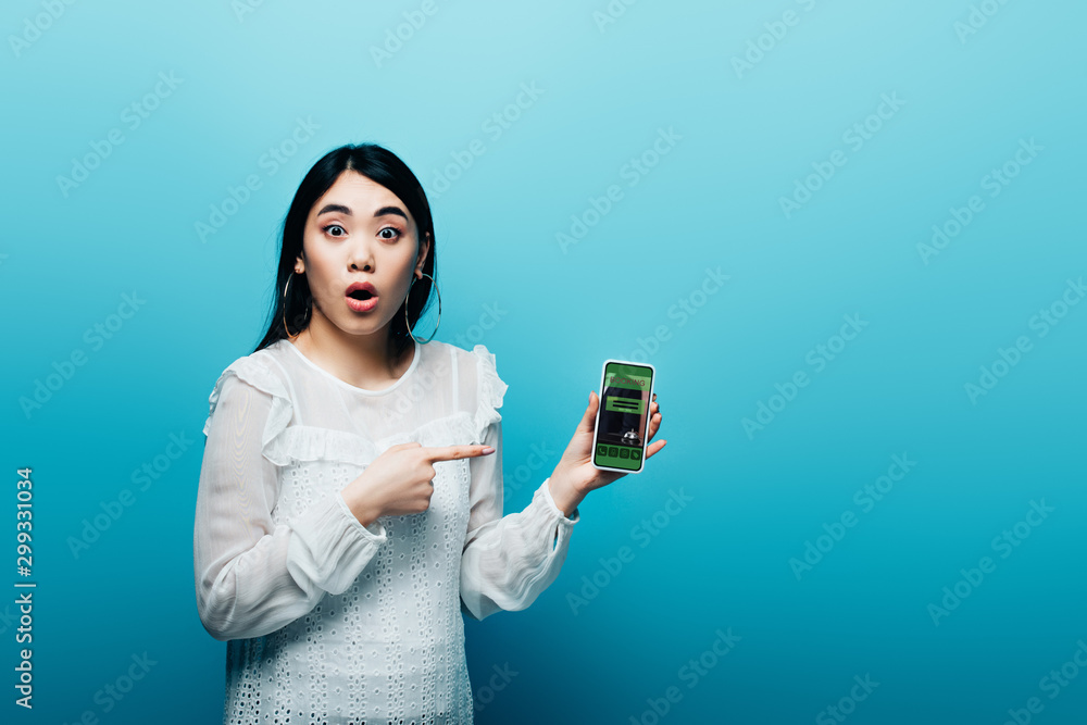 shocked asian woman pointing with finger at smartphone with booking app on blue background shocked asian woman pointing with finger at smartphone with booking app on blue background