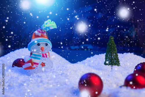 Snowman with Christmas balls on snow over fir-tree, night sky and moon. Shallow depth of field. Christmas background. Fairy tale. Macro. Artificial magic dreamy world. © Alik Mulikov