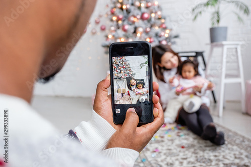 Unidentified mixed race man photographs his family blurry daughter and wife sitting on living room floor near an artificial Christmas tree in a bright interior. Concept of social networks and gadgets