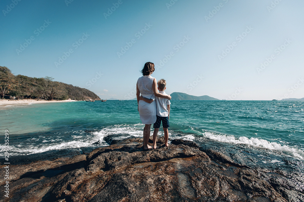 Peaceful young mother and her son hug each other and look at the turquois sea Peaceful young mother and her son hug each other and look at the turquois sea