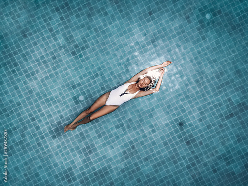 Young attractive woman floats on the back in the round blue pool, lady holds her arms up, flecks in the clear water. Luxury recreation concept, aerial view