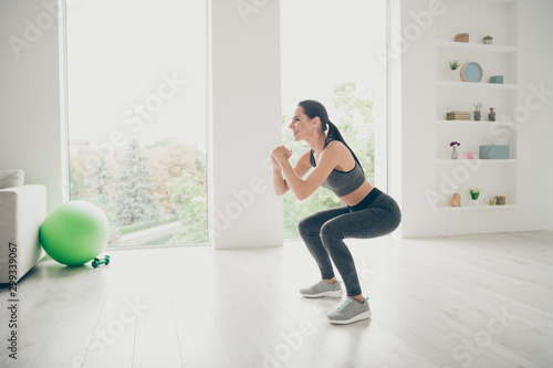 Full size profile side photo of joyful agility lover girl doing sport yoga exercises sit squats practicing pilates aerobics wear modern pants sneakers in house like fitness studio
