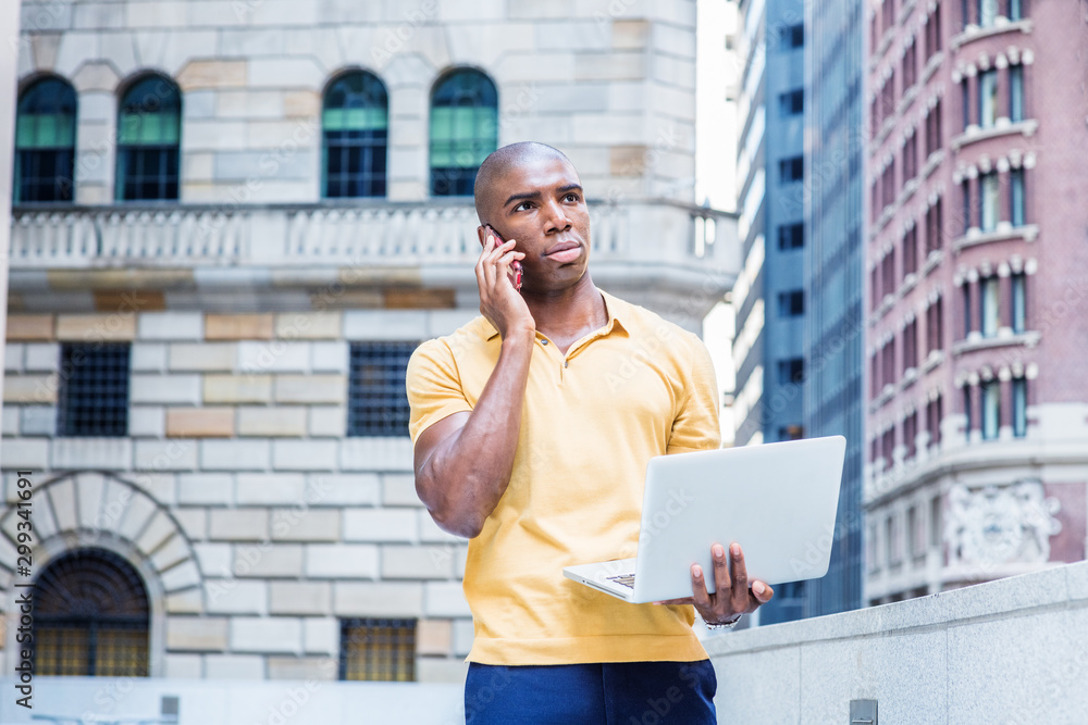 Working anywhere. Young African American Business Man working in New York City, wearing yellow short sleeve shirt, standing outside office building, talking on cell phone, working on laptop computer..