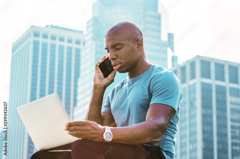 Young African American Businessman traveling, working in New York, wearing blue T shirt, sitting in front of business district, looking down, working on laptop computer, talking on cell phone. .