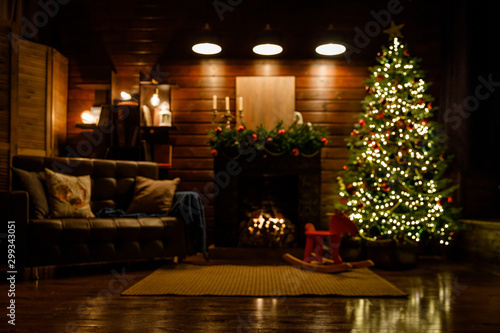Christmas and New Year interior - blur background  fireplace  lamps  green Christmas tree  brown leather sofa  gifts  candles  moose rocking chair.  Lots of lights glowing in the dark.