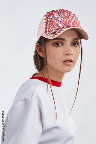 Medium close-up shot of a young European lady with straight brown hair in a white sweatshirt with red collar and a pink baseball cap adorned with pink rhinestone studding. photo