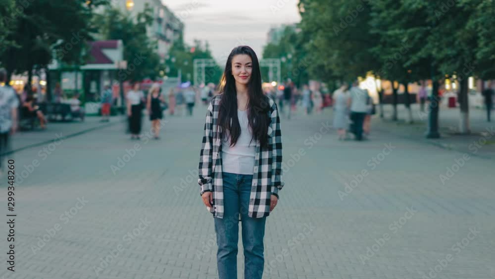 Zoom-out timelapse of pretty young lady student smiling in pedestrian street big city looking at camera standing outdoors while people are moving around Zoom-out timelapse of pretty young lady student smiling in pedestrian street big city looking at camera standing outdoors while people are moving around