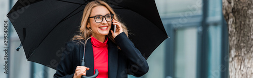 panoramic shot of attractive and smiling businesswoman in black coat holding umbrella and talking on smartphone