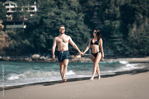 Loving couple on honeymoon walks along the sunny exotic beach holding hands, sunglasses and swimwear on; romance concept.