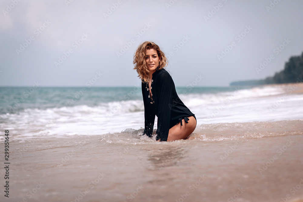 Gorgeous woman with thick blond wavy hair, wearing black long shirt sits in the water.