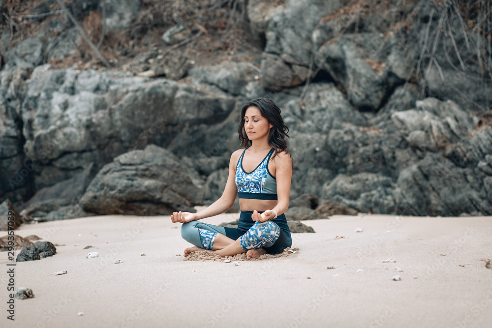 Happy slim lady meditates in lotus pose at the sandy beach by the rocks, yoga concept