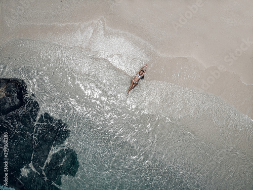 Aerial view of the fit girl lying on sandy beach by turquoise ocean waters; drone shot.