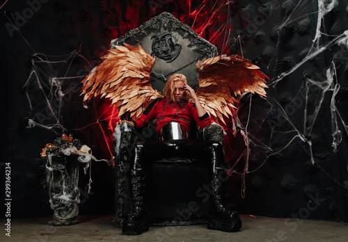 passionate lazy Cupid blond long hair guy in red leather jacket and vinyl pants with huge Golden wings sits on a huge throne in hell