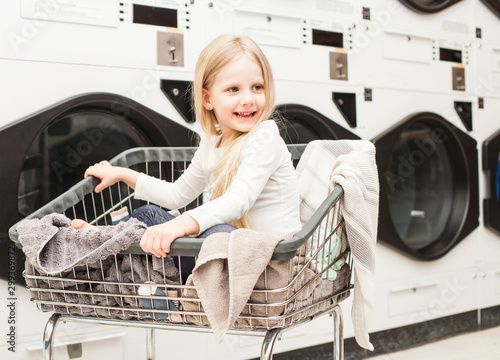 Portrait of smiling little girl sitting in cart at laundry. Happy kid posing at washing machines. Housework and hygiene