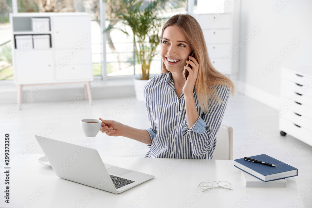 Young businesswoman talking on phone while using laptop at table in office Young businesswoman talking on phone while using laptop at table in office
