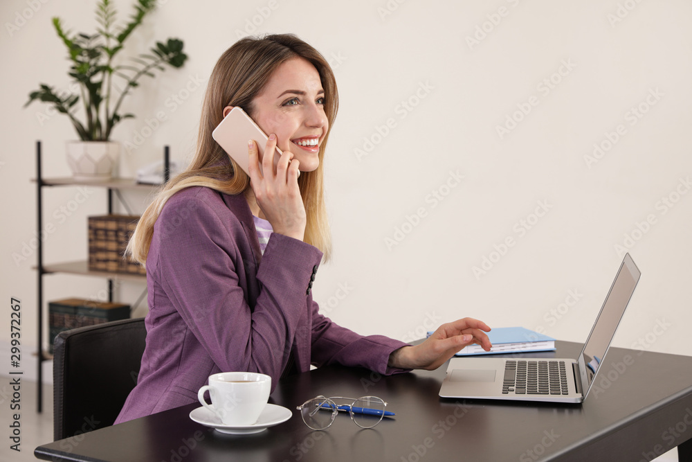 Young businesswoman talking on phone while using laptop at table in office Young businesswoman talking on phone while using laptop at table in office