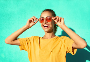 Fashionable smiling young woman in trendy red eyeglasses posing on blue background, wearing yellow t-short.