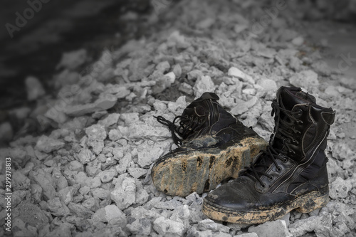 Empty ragged black combat or jungle shoes put on the construction material scraps. Old and muddy tactical soldier footwear dumped in abandoned place. Selective focus and copy space.