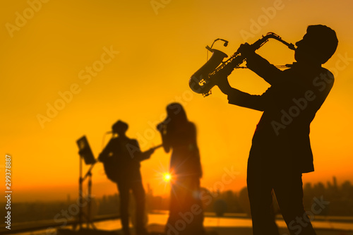 Silhouette autumn or winter scene of saxophone musician man showing with blurry jazz trio band and twilight or sunset cityscape background. Image for happy new year party or celebration concept.