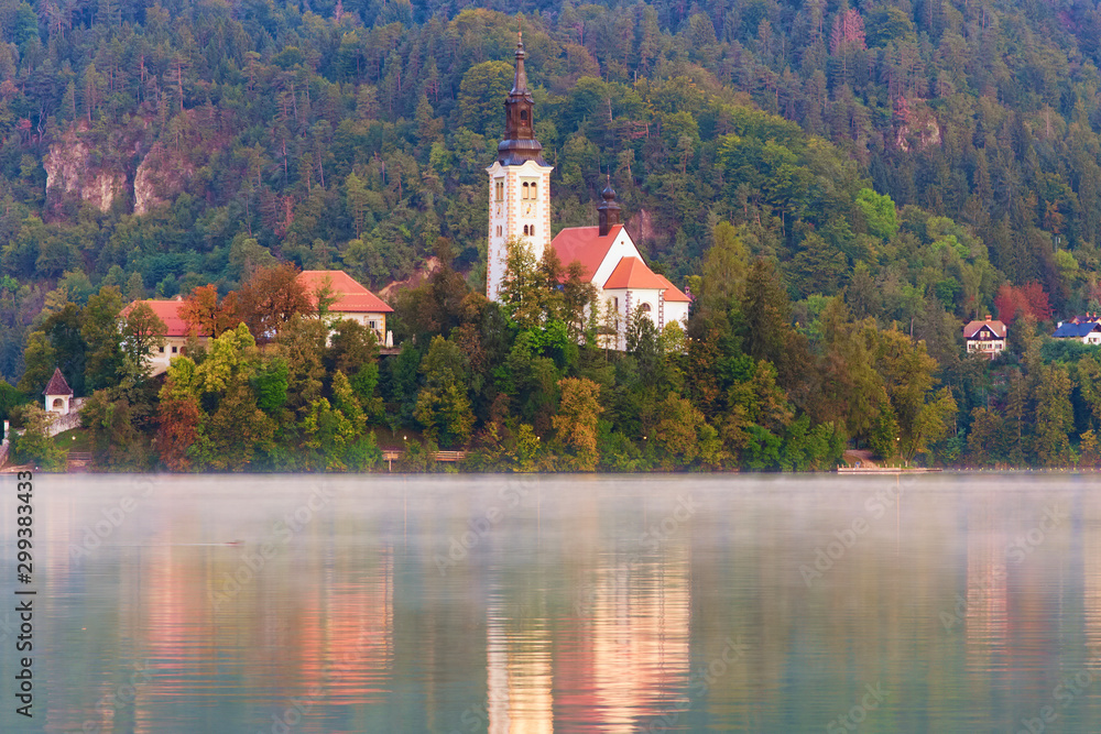 Picturesque morning view of Bled Lake and Pilgrimage Church of the Assumption of Maria on small island against autumn forest. Morning fog on the water surface. Travel and tourism concept. Slovenia