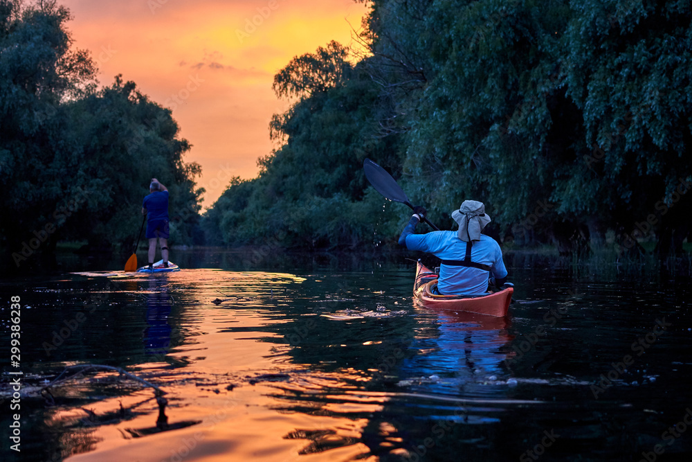 Two men paddling on SUP (stand up paddle board, paddleboard) and kayak in Danube river against the backdrop of a colorful sunset and forest along riverside