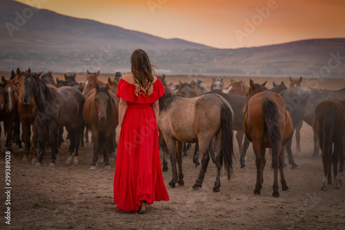 Back view of girl in red dress among many horses in sunset.