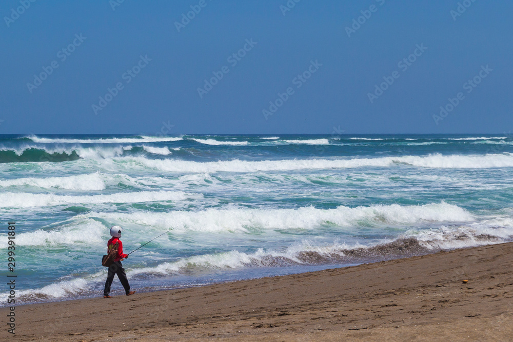 Fisherman in helmet looking like astronaut standing and fishing on the beach next to the sea