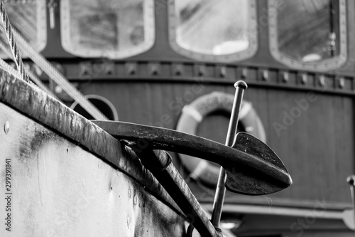 Black and white close view of an old fishing boat anchor with boat windows and lifebuoy in the background