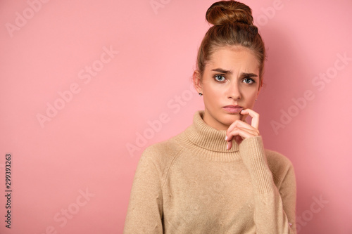 Close portrait a girl with collected hair, standing on a pink background in a beige turtleneck and frowning looking at the frame