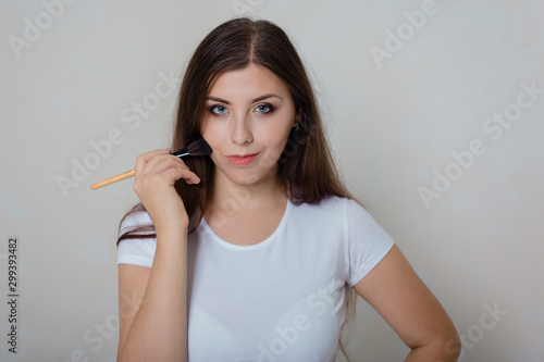 young, beautiful girl in white t-shirt doing make-up on face with cosmetic brush. Studio photo on a white photo.