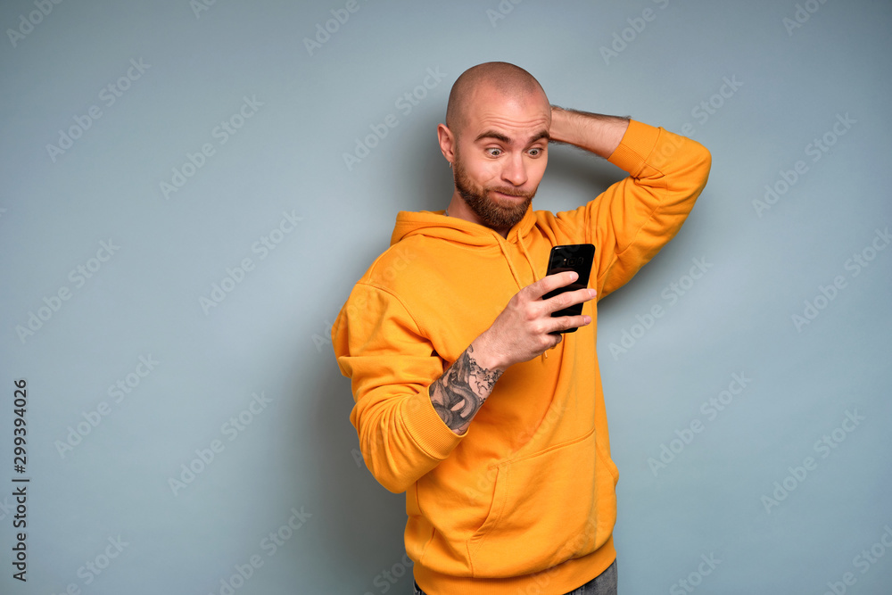 A bearded guy in a yellow sweatshirt, opening his eyes wide and pursing his lips, looks into a smartphone scratching his head A bearded guy in a yellow sweatshirt, opening his eyes wide and pursing his lips, looks into a smartphone scratching his head