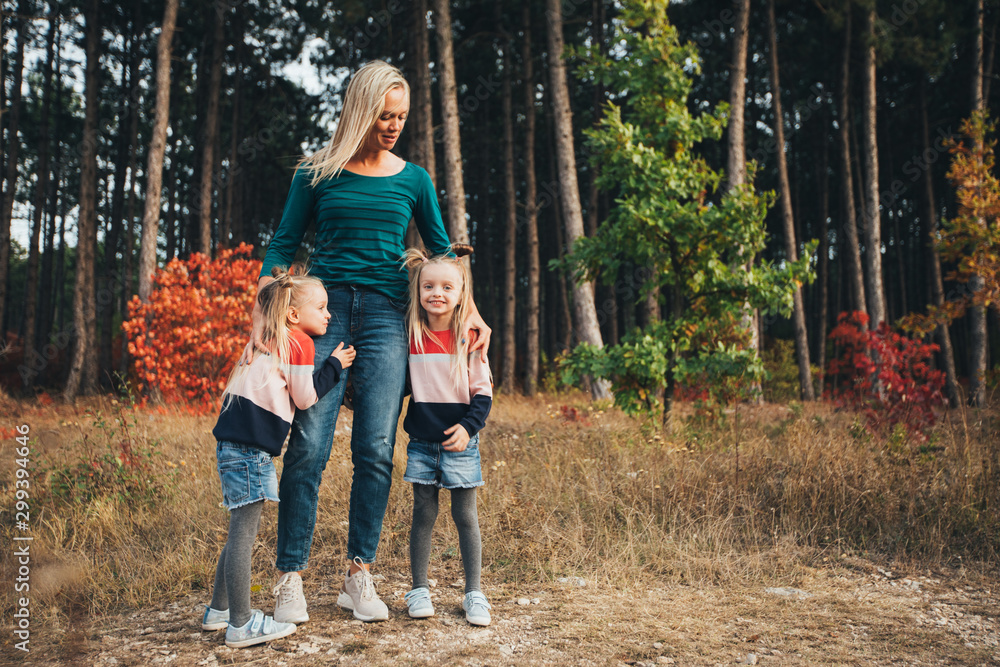 Fototapeta premium Blonde mother and her twin daughters are walking in the autumn forest among red trees.
