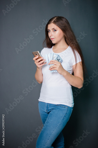 young, beautiful girl in a white T-shirt and blue jeans with a smartphone and dollars in hands. Studio photo on a gray background.